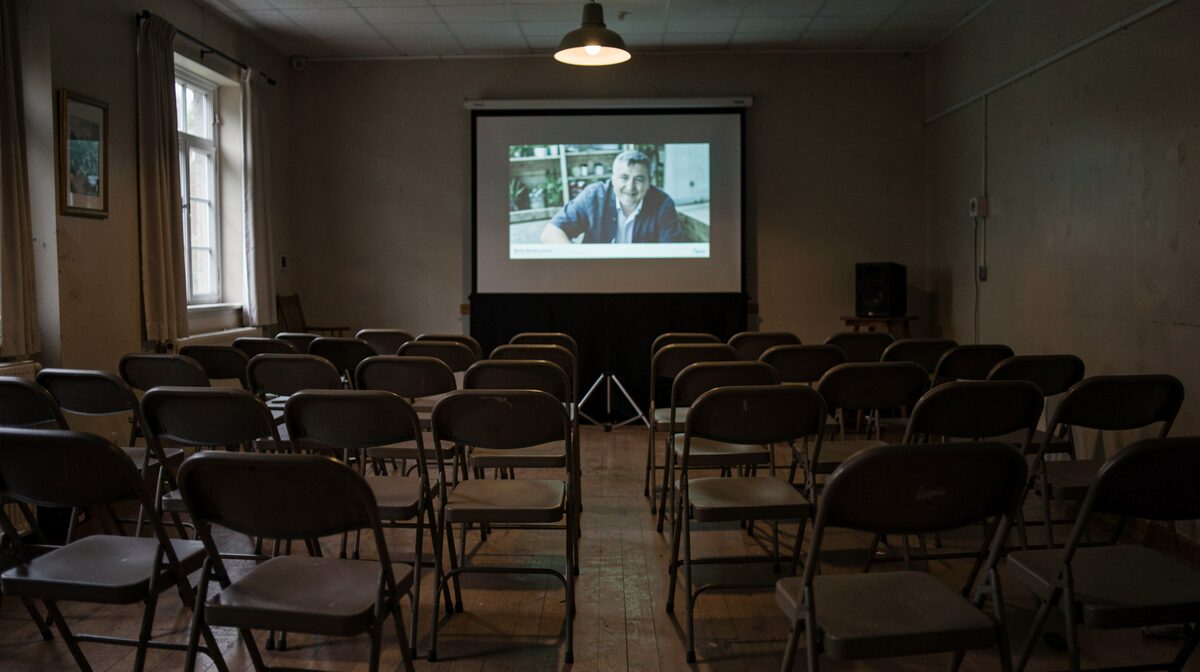 A dim community hall in St. Catharines after a screening, folding chairs in loose rows, a portable screen with a documentary still projected on it, one overhead lamp left on, framed tightly on the empty front row.